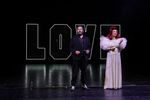 Ein Mann im Anzug und Cowboy-Stiefeln steht neben einer Frau im silbernen, extravaganten Outfit auf der Bühne. Im Hintergrund sieht man den Schriftzug "LOVE".