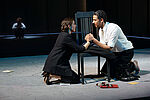 Eine Schauspielerin udn ein Schauspieler halten knieend die Hände über einem Stuhl gekreuzt. Im Hintergrund sitzt ein Schauspieler im Schneidersitz und beobachtet das Paar im Vordergrund. 