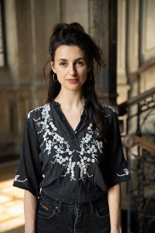 Eine junge Frau steht in einem Treppenhaus. Sie trägt eine schwarze Bluse mit weißen Verzierungen und schaut geradewegs in die Kamera.