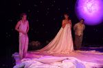 Vor einem lila Mond steht eine Frau und blickt zu zwei Männern. Der eine Mann steht in ein weißes Kleid gewickelt und hat den Mund aufgerissen. Der andere Mann trägt ein weißes Unterhemd und eine lockere Hose, an der er seine Hände hat.