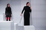 Eine Frau im schwarzen Outfit schaut hinter einen Vorhang. Hinter ihr steht eine weitere Frau mit schwarzem Rock und roter Strumpfhose. Vor den beiden sieht man weiße Blöcke stehen.