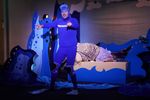 Ein Mann im blauen Neoprenanzug und Taucherflossen zeigt mit beiden Händen ins Publikum. Im Hintergrund sind Bühnenbildelemente in Wellenform zu sehen. 