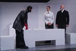 Ein Mann im schwarzen Outfit und roten Handschuhen steht an der Kopfseite eines weißen Tisches und zeigt mit einem roten Stift auf etwas, das auf dem Tisch liegt. Er trägt eine Maske über seinem Kopf, aus der nur seine Ohren und sein Gesicht rausschauen. Auf der anderen Seite des Tisches stehen eine Frau und ein Mann.
