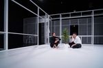 Eine Frau und ein Mann sitzen auf dem Boden, zwischen ihnen steht eine Plastikpflanze. Der Mann ist etwas.