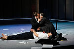 Ein Mann und eine Frau knieen auf der Bühne in einem Kuss, vor ihren Füßen liegt ausgestreckt ein gefesselter Mann.