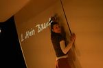 Eine Frau steht an einer Wand und hält ein Mikrofon wie einen Stift in ihrer Hand. An der Wand sind die handschriftlichen Worte "1. Mein Bruder" zu sehen.
