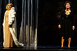Eine Frau im goldenen Kleid und Pelzmantel gekleidet steht auf der linken Seite der Bühne vor einem silbernen Vorhang. Auf der rechten Bühnenseite steht eine schwarzgekleidete Frau mit einem Kreuzanhänger an einer Kette um den Hals.