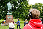 Vereinzelte Menschen mit Kopfhörern stehen vor einer Statue von Bismarck