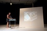Ein Mann steht an einem Tisch und hält Zettel unter eine Halterung. Auf dem Tisch steht außerdem noch ein Laptop. Vor dem Tisch liegen Fotos auf dem Boden verteilt, dahinter steht eine Leinwand, auf die ein Foto projiziert wird.