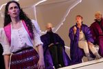Eine Frau steht links am Bildrand und blickt in die Ferne. Hinter ihr sitzen drei Männer in Roben und blicken zu ihr nach vorne.