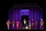 Ein Mann steht im Glitzerjacket mit einem Mikrofon auf der Bühne. Hinter ihm stehen acht Drag-Queens in bunten Outfits vor einem lilafarbenen Vorhang.