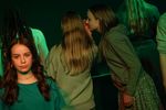 Eine Jugendliche hat sich zu einer weiteren geneigt, die mit dem Rücken zur Kamera steht, und flüstert ihr etwas ins Ohr. Neben den beiden stehen weitere Jugendliche mit dem Rücken zur Kamera.