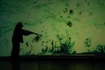 Vor einer grüngefleckten Projektion sieht man die schwarzen Umrisse einer Person, die ein Gewehr in der Hand hält.