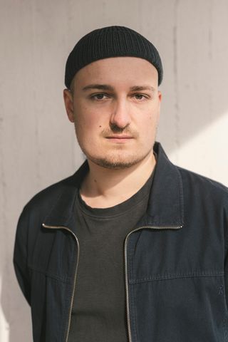 Ein junger Mann steht vor einer weißen Wand, auf die ein Lichtstrahl fällt. Der Mann trägt eine Mütze und hat dunkle Oberteile an.