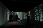 Ein Mann steht vorne im Bild und knüpft sein Hemd. Hinter ihm sieht man drei weitere Personen in der Ecke stehen. Auf die Wände hinter den Personen sieht man die Projektion eines Waldes.
