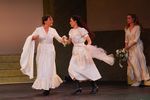 Zwei Frauen in weißen Kleidern laufen Hände haltend. Hinter den beiden steht eine weitere Frau mit Blumenstrauß in der Hand und weißem Kleid.