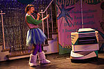 Elisabeth Frank als Jurrit in einem bunten Kostüm, mi dem Keksobil, vor einer Neonanzeige und einem Glitzervorhang