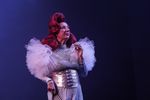 Eine Frau mit roten Haaren hält eine Pfeife zwischen ihren Fingern. Sie trägt ein extravagantes silbernes Outfit mit Rüschen an den Schultern.