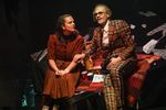 Eine Frau in einem Kleid sitzt auf einem Podest, neben ihr ein Mann im karierten Anzug und schwarzer Brille. Zwischen den beiden steht ein Picknickkorb mit Kanne und Tassen, sowie eine Zeitschrift.