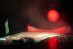 Unter einem roten Mond liegt eine riesige weiße Decke, die von zwei Menschen gehalten wird. Ein dritter Mensch steht neben der Decke.