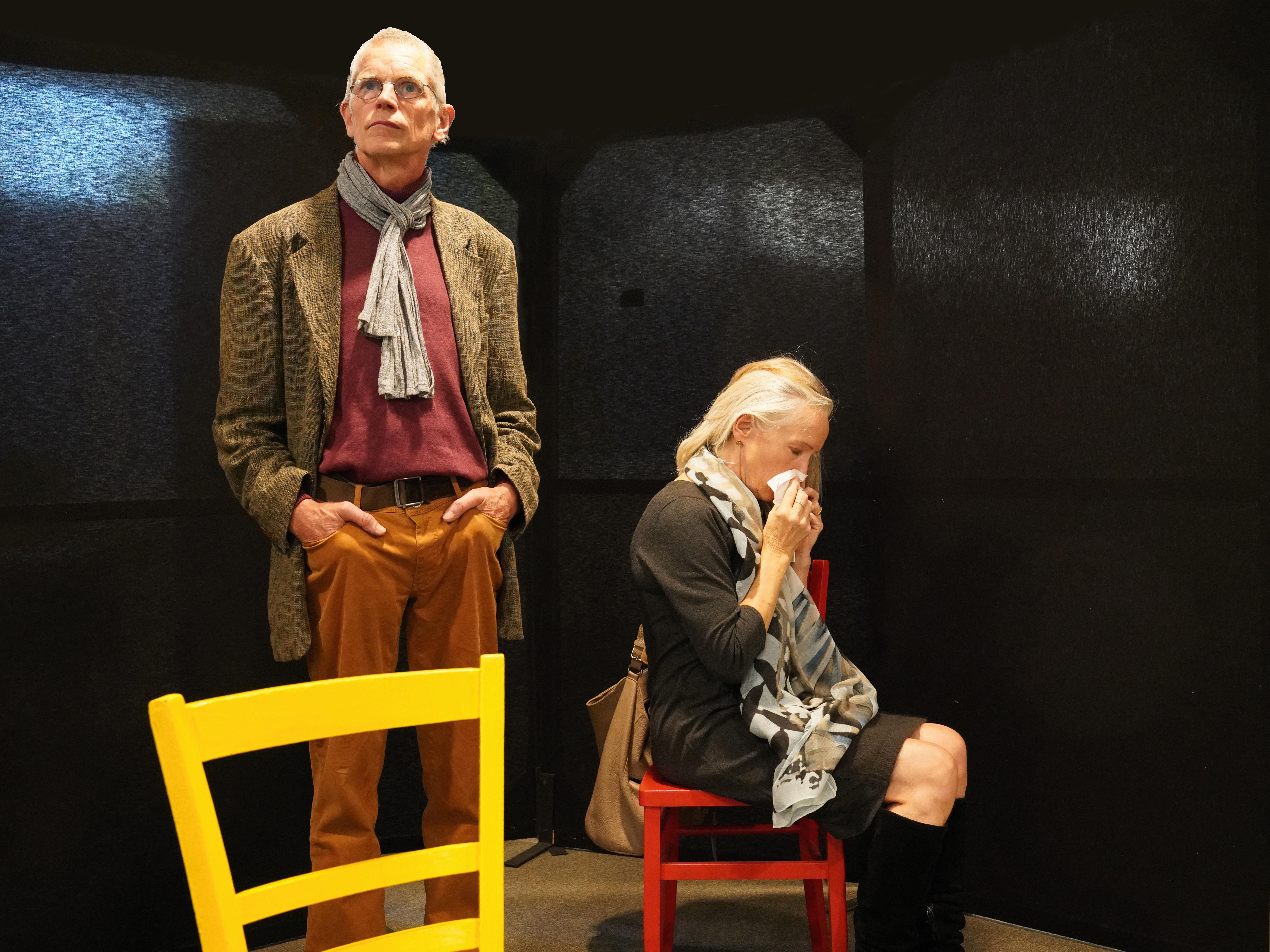 Eine Frau sitzt auf einem roten Stuhl und schnäuzt sich die Nase. Links neben ihr steht ein Mann hinter einem gelben Stuhl und hat die Hände in die Hosentaschen gesteckt.